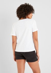 Woman with curly hair facing away wearing a white short-sleeve shirt with black trim and black rolled-up shorts against a plain background.