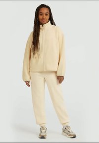 Cream-colored fleece jacket with a high collar and full zipper, paired with matching fleece pants. Smooth texture, relaxed fit.