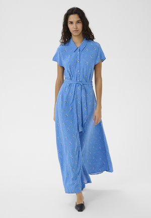 Kobieta ubrana w niebieską, kwiecistą, maxi sukienkę na guziki z krótkim rękawem, z paskiem przewiązanym w talii i czarne buty wsuwane, stojąca na jednolitym tle.