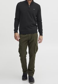 Pull noir à fermeture éclair avec poignets et ourlet côtelés, associé à un pantalon cargo olive avec poches latérales, porté avec des bottes noires à lacets.