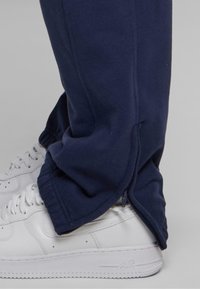 Marineblaue Jogginghosen mit glatter Textur, ausgestattet mit seitlichen Reißverschlüssen an den Knöcheln, kombiniert mit weißen Sneakers.