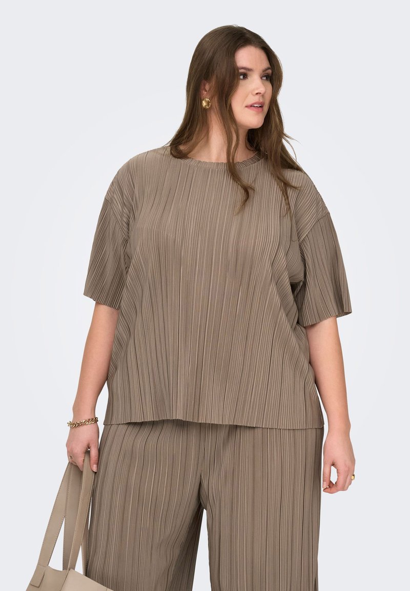 Γυναίκα που φοράει χαλαρή τοπ σε ταμπά χρώμα με πλισέ λεπτομέρειες και ταυτόχρονο φαρδύ παντελόνι, κρατώντας μπεζ τσάντα χειρός, κοιτάζοντας δεξιά με μακριά καστανά μαλλιά και χρυσά σκουλαρίκια.