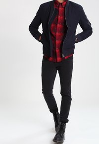 Chaqueta bomber azul marino sobre una camisa de cuadros roja, jeans negros de corte ajustado y botas negras con cordones. Cuenta con puños acanalados y cierre de cremallera.