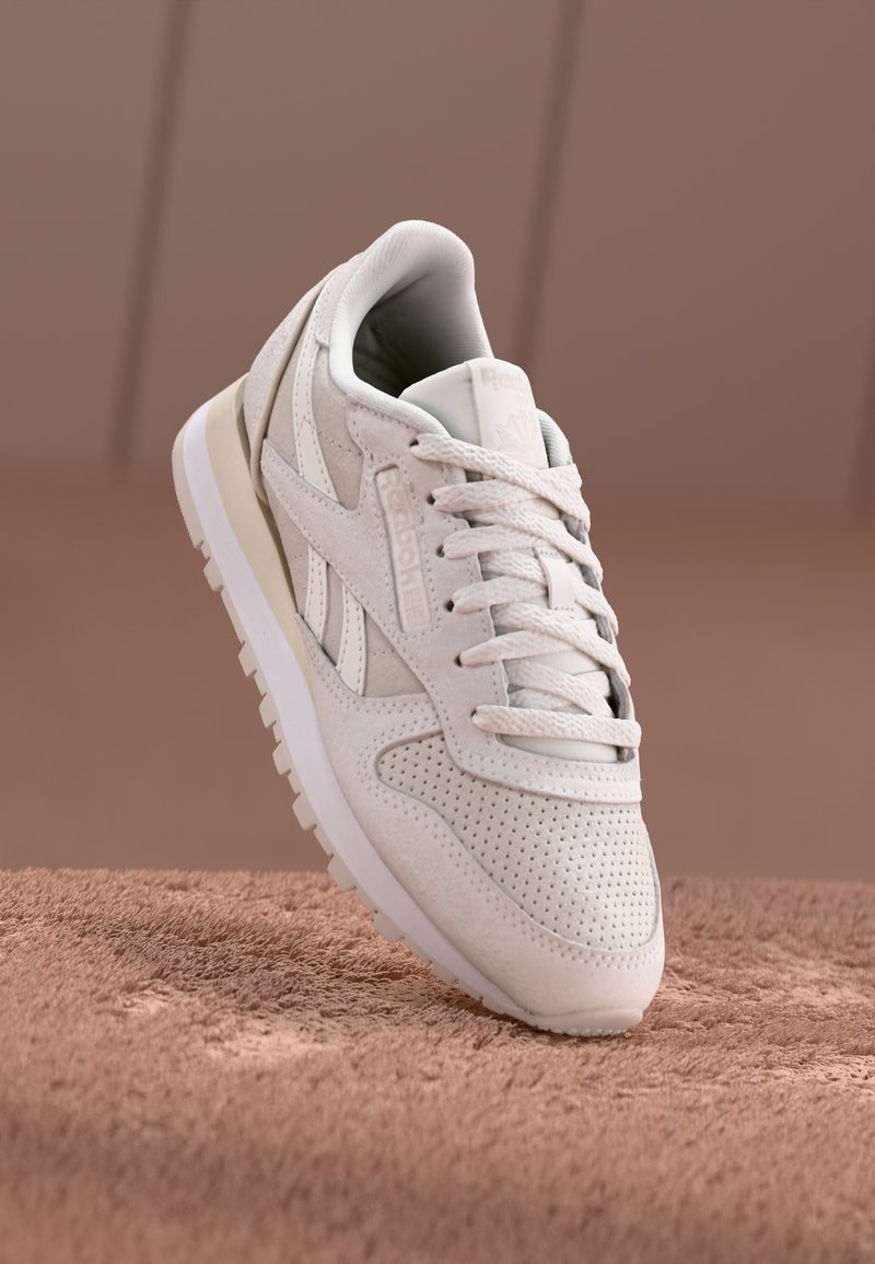 White suede athletic shoe featuring perforated detailing, flat laces, a cushioned collar, and a textured white rubber sole.