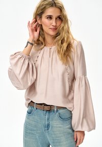 Kobieta o falowanych blond włosach, ubrana w bladoróżową bluzkę z bufkami i falbanami, niebieskie dżinsy oraz brązowy pasek.