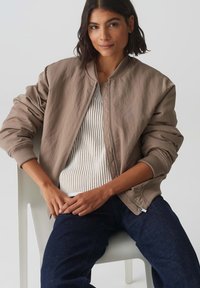 Giubbotto bomber marrone con polsini a costine, con un design trapuntato, indossato sopra una maglietta bianca a righe, abbinato a jeans blu scuro.