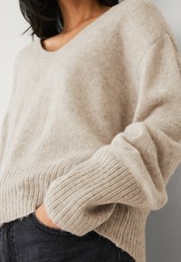 Beige gebreide trui met een V-hals, geribbelde manchetten en een losse pasvorm, met een zachte textuur en een zoom die iets omhoog loopt.