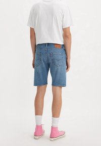 Pantalones cortos de mezclilla en azul claro con corte recto, diseño de cinco bolsillos y un parche trasero de cuero marrón. Combinados con zapatillas de deporte altas rosas.