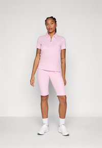 Camisa polo rosa claro com fecho zip, combinada com calções rosa claro até aos joelhos. Ténis brancos de desporto completam o visual. Textura suave.