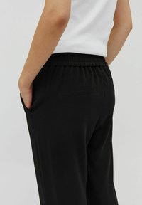 Zwarte elastische taillebandbroek met achterzakken, gladde textuur, comfortabele pasvorm en zachte stof. Model staat met hand in de zak.