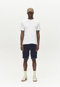 Maglietta bianca, pantaloni corti blu navy, cappellino beige con testo, accenti gialli, sneakers bianche e beige, e calzini crema, su un modello maschile contro uno sfondo semplice.