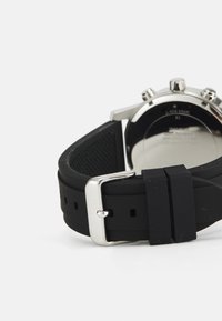 Cinturino per orologio in silicone nero con fibbia in metallo argentato, caratterizzato da una superficie testurizzata e diversi fori di regolazione, attaccato a un cassa dell'orologio in metallo.