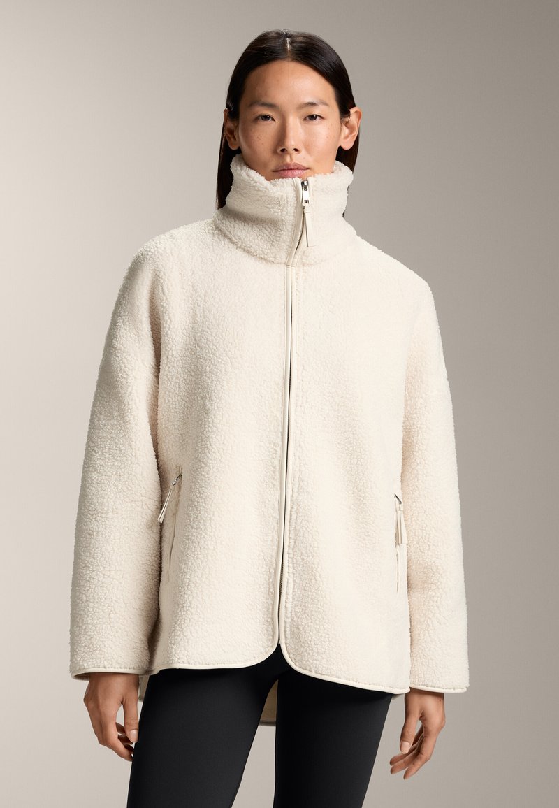 Giacca in pile crema chiaro con colletto alto e zip frontale completa. Presenta tasche laterali con zip e una vestibilità comoda, esposta su uno sfondo neutro.