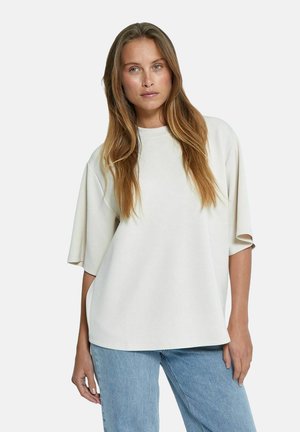 T-shirt oversize beige realizzata in tessuto morbido, con collo rotondo e maniche corte. Abbinata a jeans blu chiaro a vestibilità ampia.