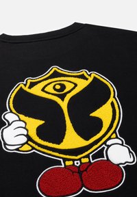 Zwarte sweatshirt met een geborduurd ontwerp met een geel embleem, zwarte accenten en rode schoenen. Textuurdetails vergroten de visuele aantrekkingskracht.