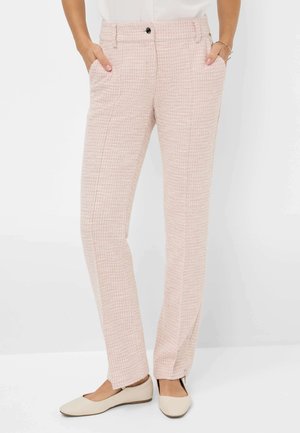 Femme portant un pantalon droit texturé rose clair avec les mains dans les poches et des chaussures plates couleur crème sur fond blanc.