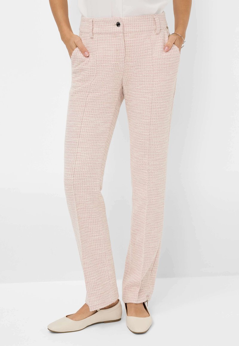 Femme portant un pantalon droit texturé rose clair avec les mains dans les poches et des chaussures plates couleur crème sur fond blanc.