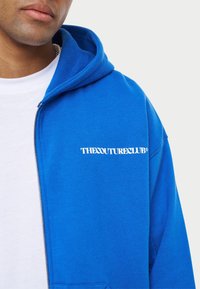 Niebieska bluza z zamkiem wykonaną z miękkiego materiału. Posiada przednią kieszeń oraz biały napis logo "THECOUTURECLUB" na lewej części klatki piersiowej.