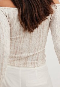 Kvinna med långt brunt hår, iklädd en off-shoulder strukturad beige topp och vita byxor, sedd bakifrån.