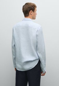 Camicia a righe azzurre chiare con orlo arrotondato. Maniche lunghe e colletto standard, realizzata in un tessuto testurizzato. Indossata con pantaloni scuri a righe.