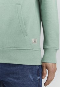 Mintgroene sweatshirt met een kangoeroezak, ribgebreide boorden en onderrand. Bevat een label aan de zijkant met tekst en een jaarindicatie.