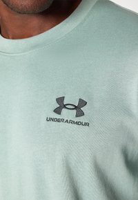 Világos zöld sportpóló, pamut keverék anyag, kerek nyakkivágás, fekete Under Armour logóval a mellkasán, sima textúra, minimalista dizájn.