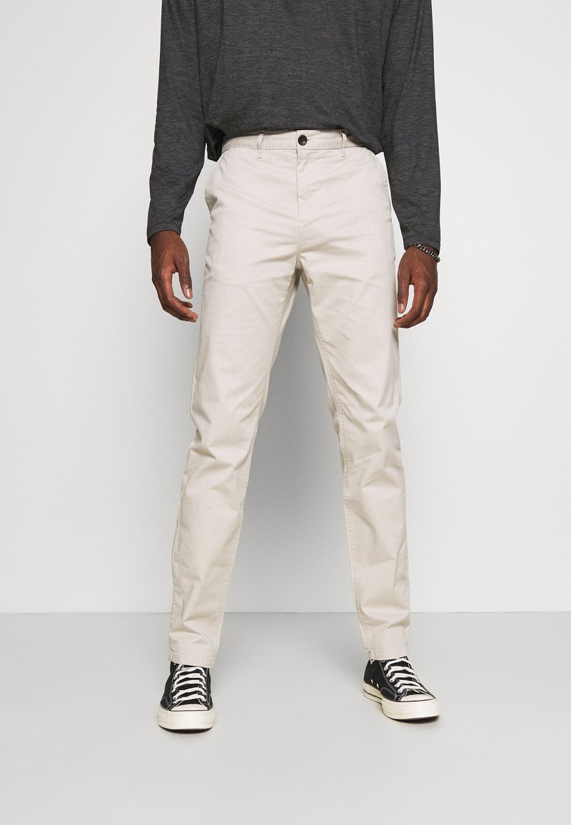 Chino in cotone color beige con vestibilità slim, dotati di chiusura a bottone e passanti per cintura, abbinati a sneakers nere con suola e lacci bianchi.