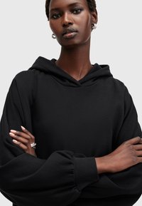 Sudadera negra con capucha y corte relajado, que presenta mangas anchas y una textura suave. Los brazos del modelo están cruzados sobre el pecho.