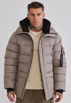 Homme avec des tatouages au cou portant une doudoune beige avec une doublure noire à la capuche, un pull beige et des colliers en chaînes argentées superposés.