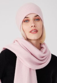Cappello a maglia rosa chiaro con texture a coste e sciarpa morbida rosa abbinata, entrambi realizzati in un materiale caldo, indossati sopra un maglione nero.
