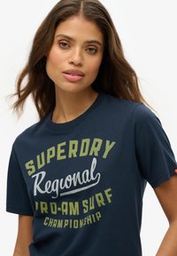 Camiseta de algodón azul marino con el texto "SUPERDRY Regional Pro-Am Surf Championship" en amarillo y blanco. Manga corta y cuello redondo.