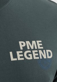 Mørkegrøn t-shirt med rund hals, der har fed hvid tekst "PME LEGEND" og detalje med blåt rundt logo. Blødt, tekstureret bomuldsstof.