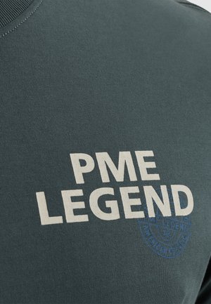 Tamnozelena majica s okruglim izrezom, s podebljanim bijelim natpisom "PME LEGEND" i plavim okruglim logotipom. Mekana, teksturirana pamučna tkanina.