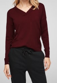 Frau trägt einen dunkelroten V-Ausschnitt-Pullover und schwarze Hose mit Kordelzug, steht vor einem schlichten, hellen Hintergrund.