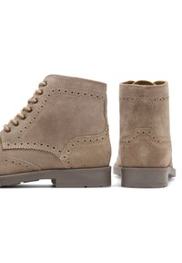 Stivaletti in camoscio marrone con dettagli brogue, punta rotonda e chiusura di tipo stringata. Suola in gomma e accenti testurizzati sulla tomaia.