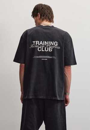Osoba stoi tyłem do kamery i ma na sobie czarny T-shirt z napisem "Training Focus on Progress Club" dużymi białymi literami.