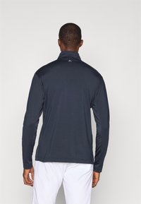 Granatowa koszulka sportowa z długim rękawem, z wysokim kołnierzem, gładką teksturą i dopasowanym krojem. Posiada małe logo na szyi oraz kontrastowe panele rękawów.