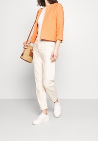 Veste orange texturée, haut côtelé rose clair, jean crème et baskets blanches. Tient un sac tissé beige avec un accent marron.