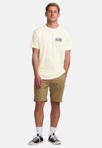 Light yellow cotton t-shirt with a front pocket graphic, paired with beige shorts, white socks, and black sneakers.