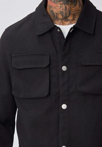 Veste noire dotée de deux poches avant à rabat, boutons-pression, et d'un col structuré net, fabriquée en tissu résistant.