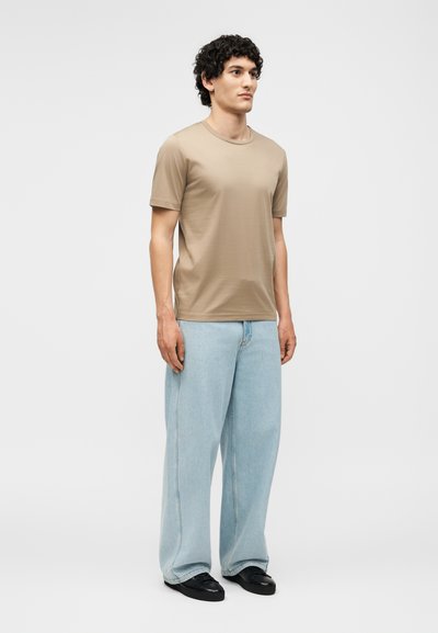 Άνδρας στέκεται φορώντας μπεζ κοντομάνικη μπλούζα, ανοιχτό μπλε φαρδιά τζιν παντελόνια και μαύρα παπούτσια μπροστά σε απλό ανοιχτό φόντο.