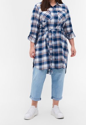 Vrouw die een lange blauw-wit geblokte blouse draagt met opgerolde mouwen, lichtblauwe cropped jeans en witte sneakers, staand tegen een witte achtergrond.