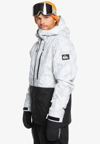Weiße und schwarze Schneejacke mit einem Camouflage-Muster, ausgestattet mit einem hohen Kragen, verstellbarem Kapuze und Klettverschlussärmeln. Inklusive Handschuhe und Goggles.