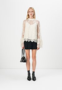 Blusa bianca in pizzo con maniche lunghe trasparenti e motivi floreali, indossata sopra una gonna nera plissè, abbinata a calze scure e tacchi neri.