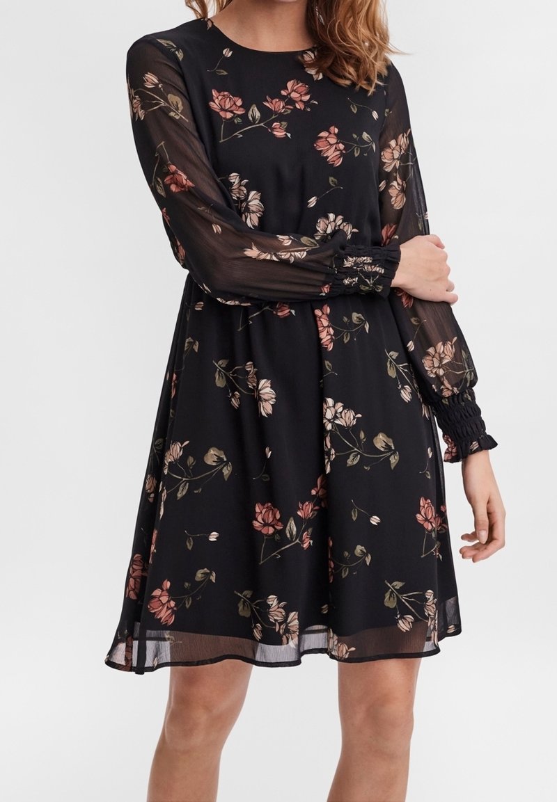 Robe noire longueur genoux à motif floral rose, manches longues transparentes avec poignets smockés, portée par une femme les bras croisés.
