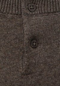 Prim-plan al unei țesături tricotate maro cu două butoane întunecate și o mică buzunar rectangular pe partea stangă.