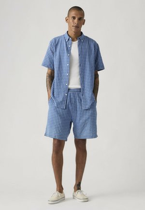 Mężczyzna z krótkimi włosami, ubrany w niebieską koszulę w kratę z krótkim rękawem i dopasowane spodenki, na białym podkoszulku, stoi z rękami w kieszeniach.