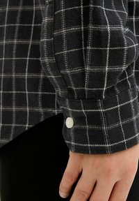 Melna un pelēka svītrainā flaneļa krekla piedurkne, kurai ir balta šuve un pogu slēgšana. Mīksta tekstūra ar brīvu piegriezumu un pielāgotām šuvēm.