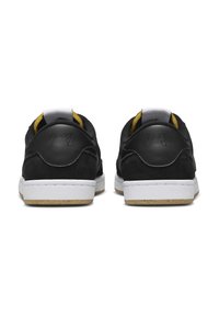 Musta mokkanahkainen tennari, jossa on valkoinen kumipohja, keltaisia yksityiskohtia ja musta kantapään nauhanlogo kohokuvioidulla yksityiskohdalla.