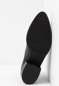 Semelle de botte en cuir noir à bout pointu avec une surface en caoutchouc texturée, reposant contre un mur et une surface blancs.